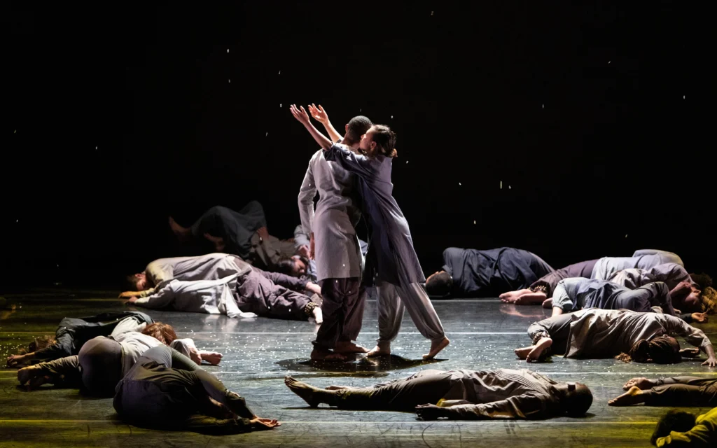 A dozen dancers lie still on the ground, huddled on their sides and sprawled on their backs. At the center, two dancers stand close together, facing each other. One reaches their arms up at an angle around the other's shoulders, as they twist so they face upstage. Snow falls.