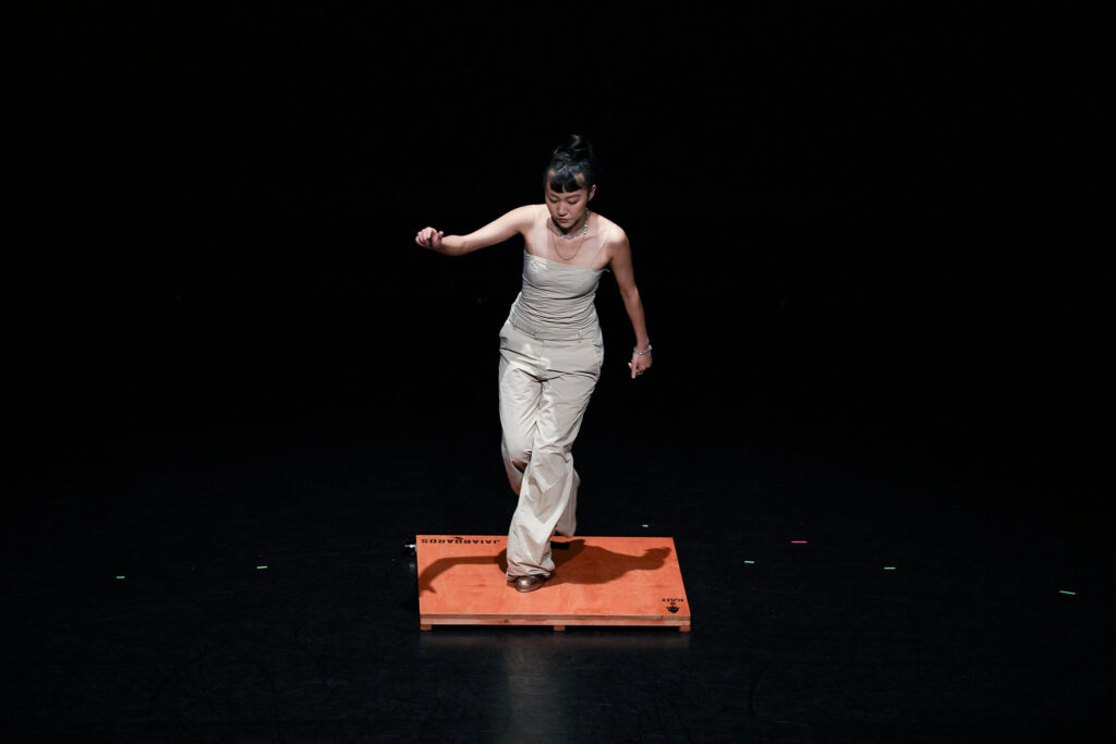 A woman in a white top and pants stands on a wooden platform, stepping forward one arm raised