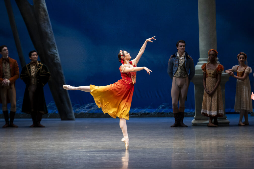 a female dancer in third arabesque on stage wearing an orange and yellow dress 