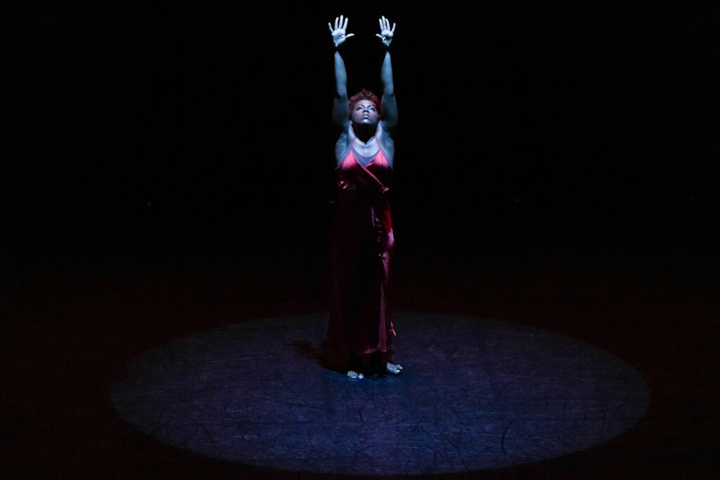 A woman stands in a long red dress, her arms raised over her head