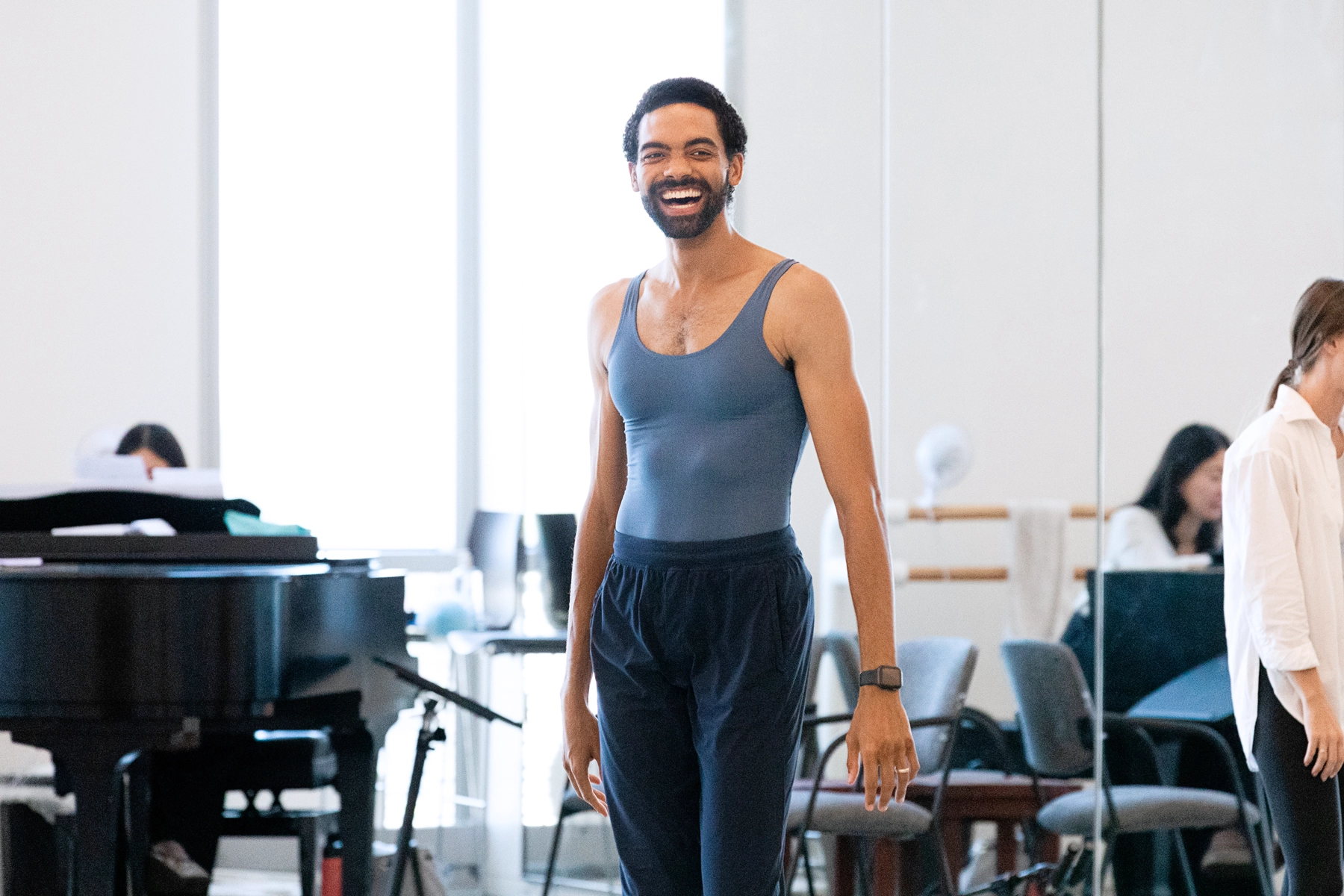 Silas Farley, a tall, lanky Black man, smiles brightly at the front of a studio. He wears a fitted blue biketard and dark sweatpants.