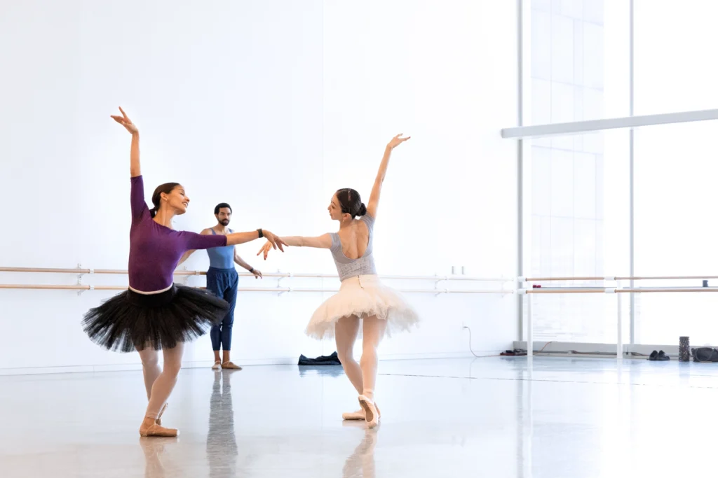 In a bright, airy ballet studio, two dancers in practice tutus and pointe shoes pose in tendu fondu back, facing opposite directions. They arch toward each other as they look over their shoulders toward center. Silas Farley watches from the back of the studio.