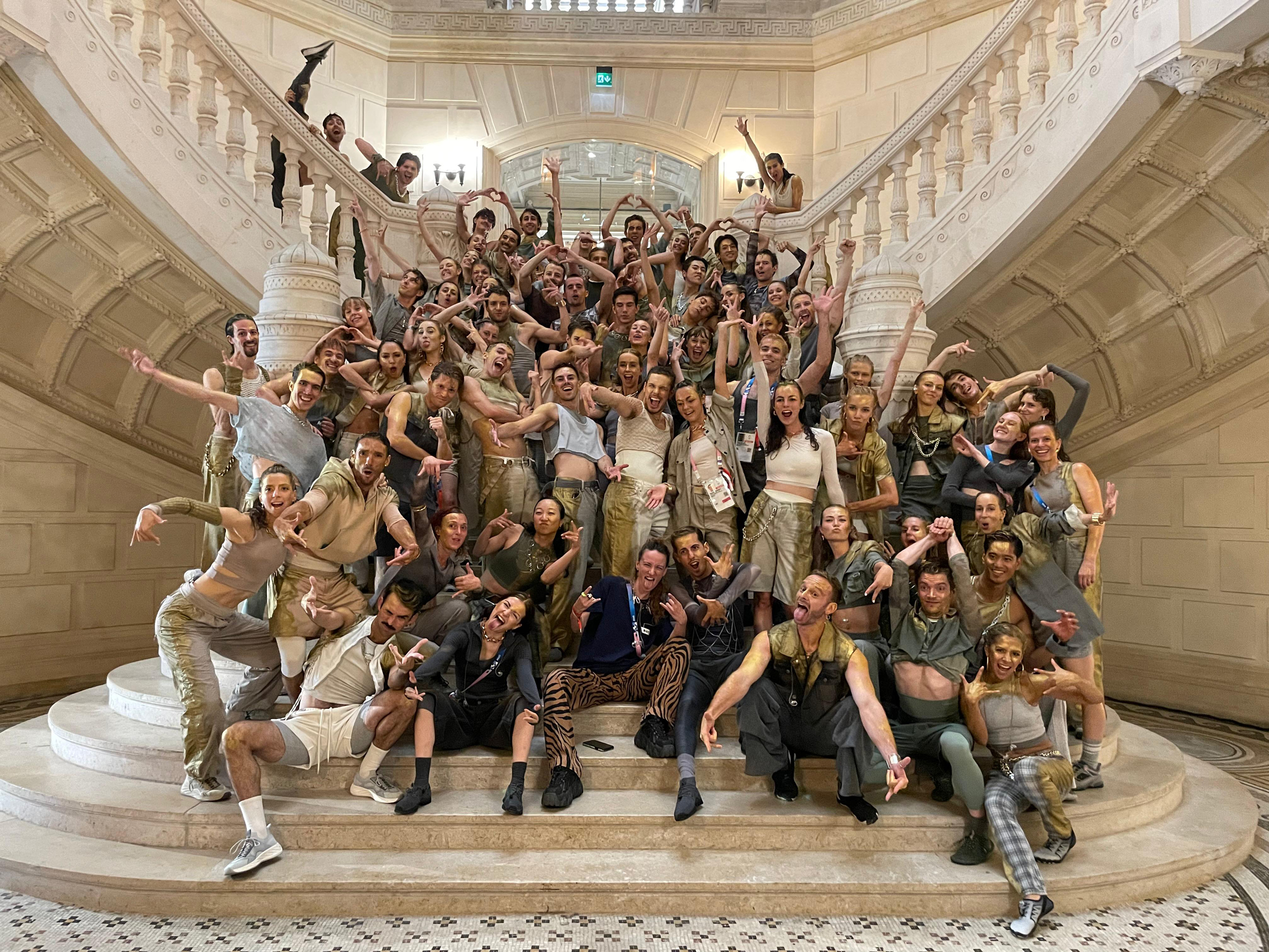 A large group of dancers in grey and gold costumes strike exuberant poses on a massive, intricately carved marble staircase.