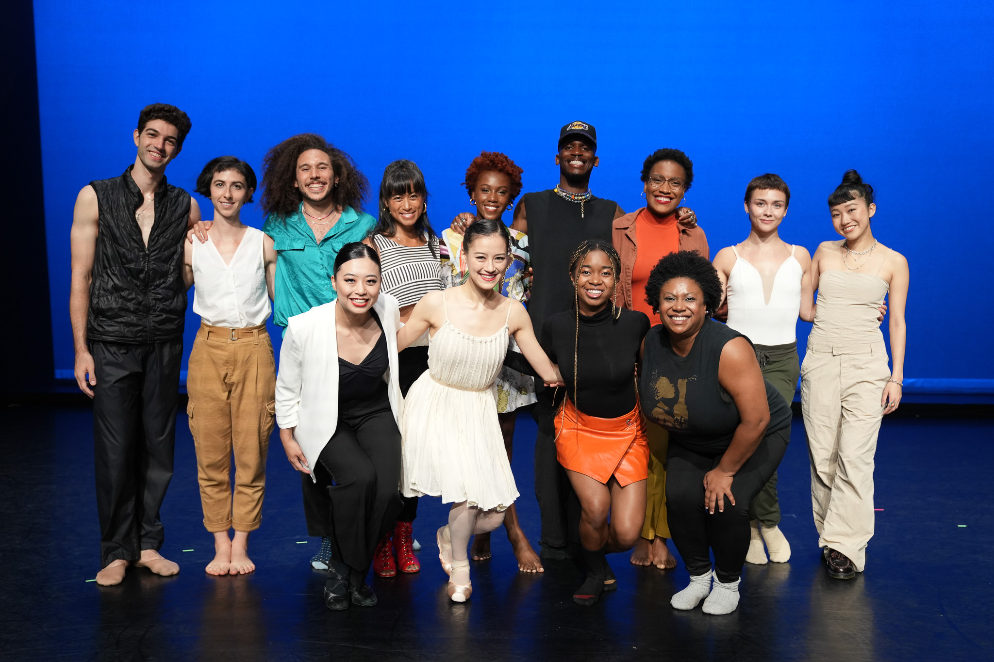 A group of 13 artists stand in a group onstage in two rows in front of a blue background