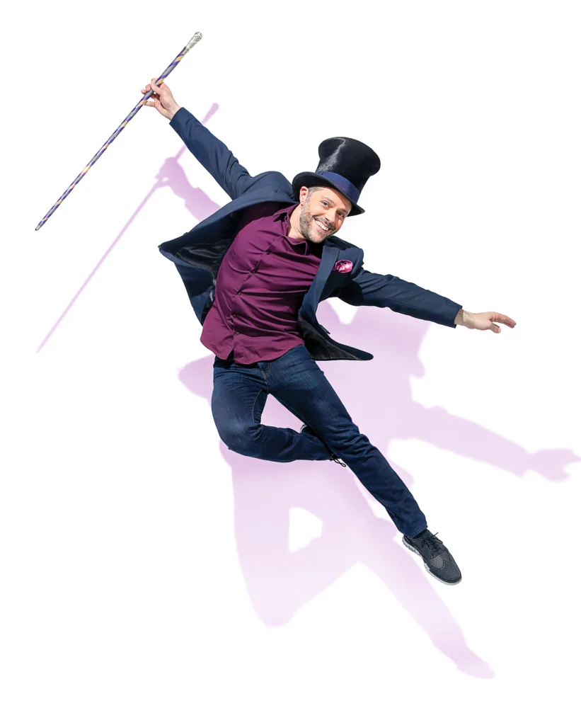 Joshua Bergasse, wearing a suit with a plum colored shirt and a top hat, leaps against a white background, casting a shadow behind him. He holds a walking stick in one hand, one foot tucked behind the knee of a straight bottom leg.