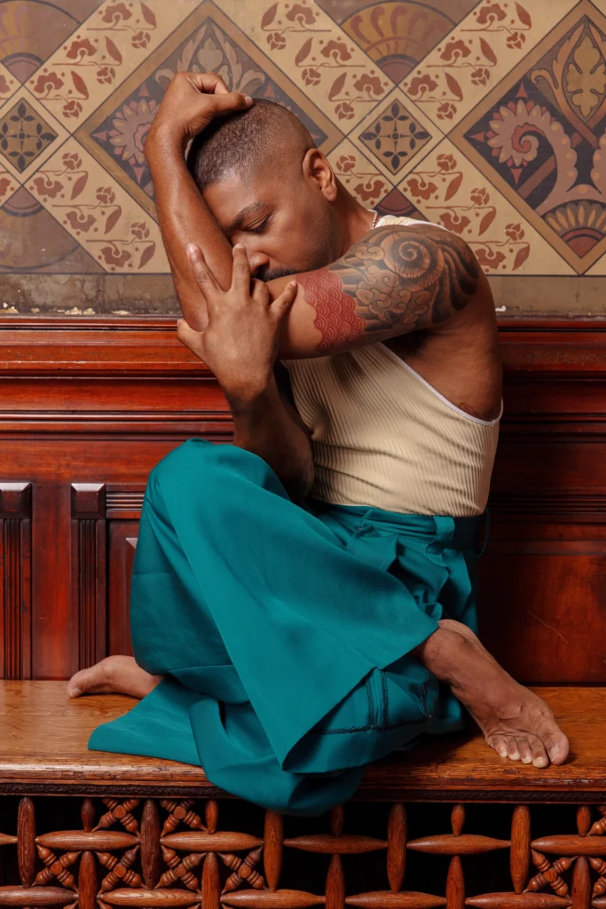 Kyle Abraham sits on a wooden bench against a richly patterned wall. His legs are crossed beneath him, arms wrapping around his chest to cradle his head. He wears teal pants and a white tank.