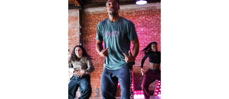 Carr, a Black man with a small goatee wearing a green t-shirt, jeans, and gold hoop earrings, is caught mid-gesture as he leads a class of dancers in a brick-walled studio.