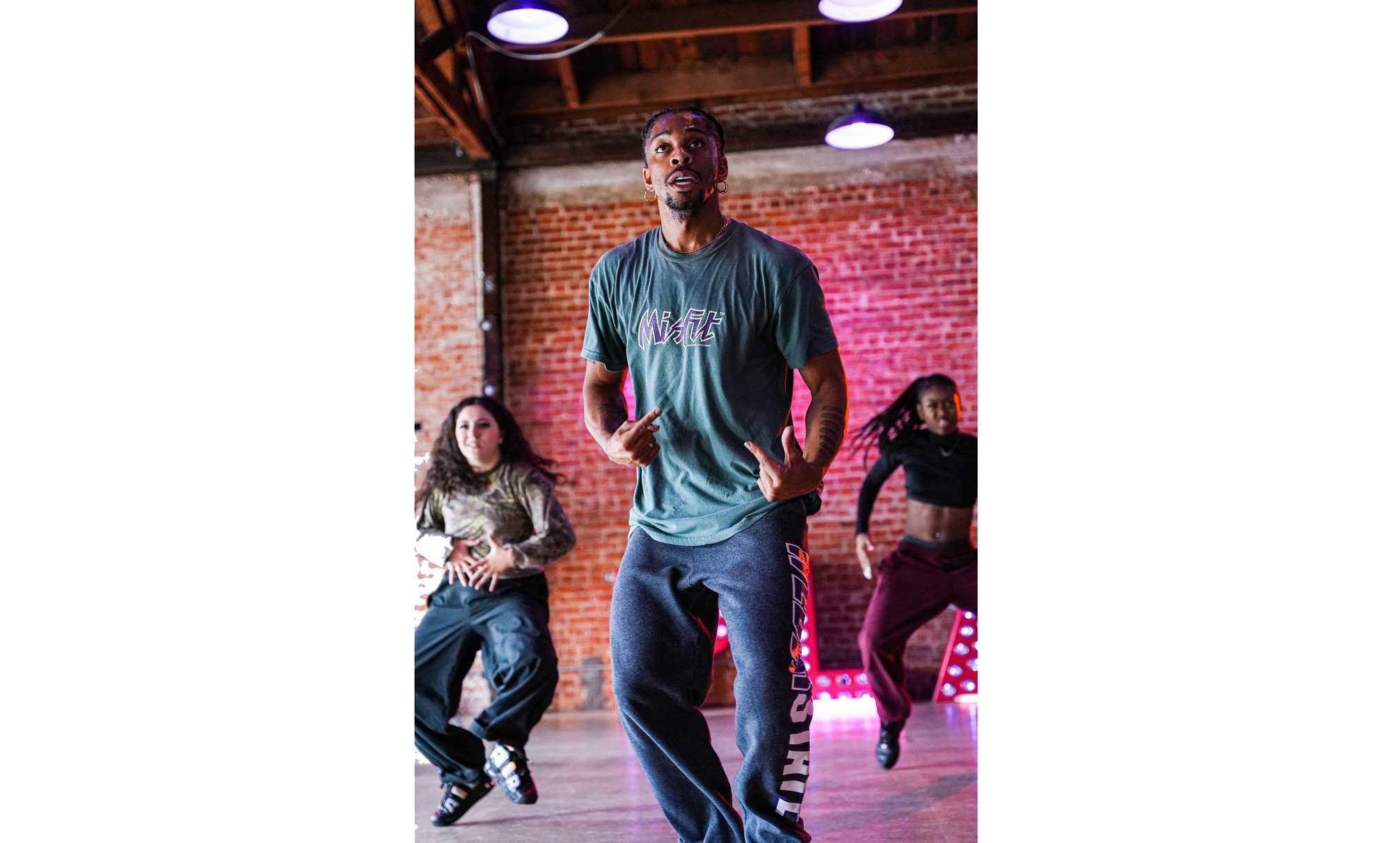 Carr, a Black man with a small goatee wearing a green t-shirt, jeans, and gold hoop earrings, is caught mid-gesture as he leads a class of dancers in a brick-walled studio.