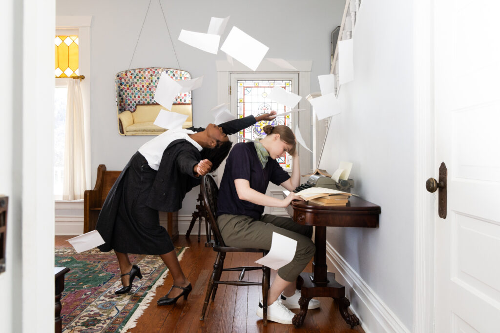 two dancers in a house, one sitting at a desk reading with the other arching back and throwing papers into the air