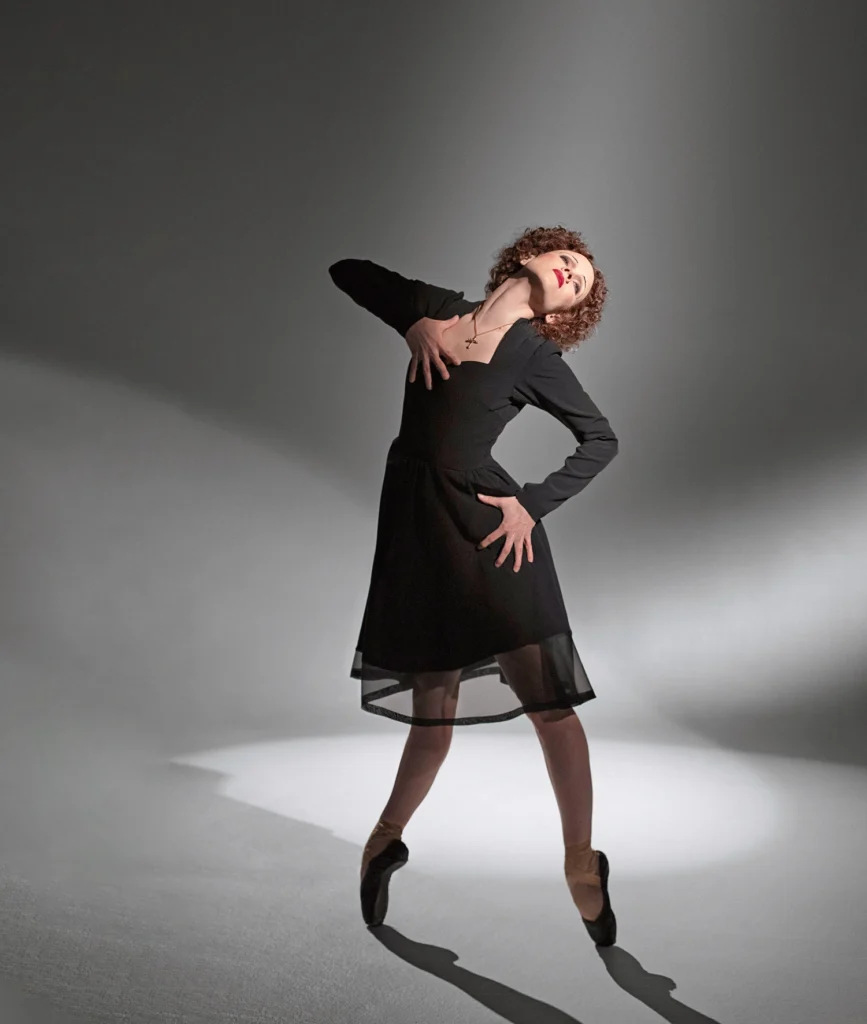 A dancer in a structured black dress and pointe shoes poses in front of a spotlight. She presses one palm to her hip, the other to her chest, as she arches back and tips her head toward the light.