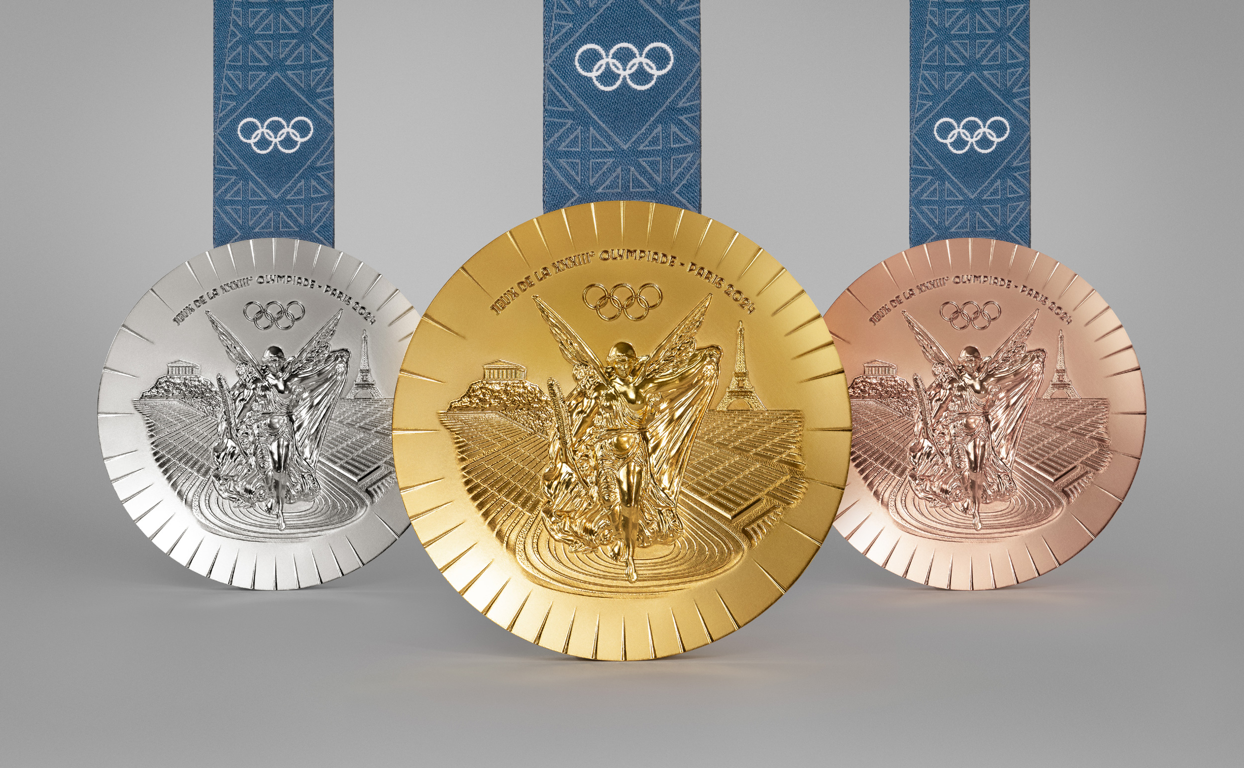 A close-up image of finely detailed silver, gold, and bronze medals on a plain gray background.