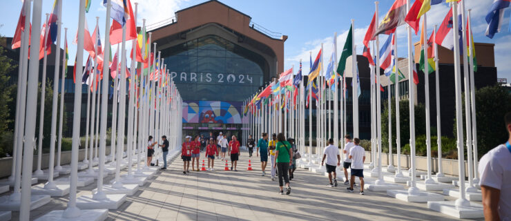 Scattered groups of people walk down a wide path lined with flags of many nations and leading to a large glass-walled building with a "Paris 2024" sign.