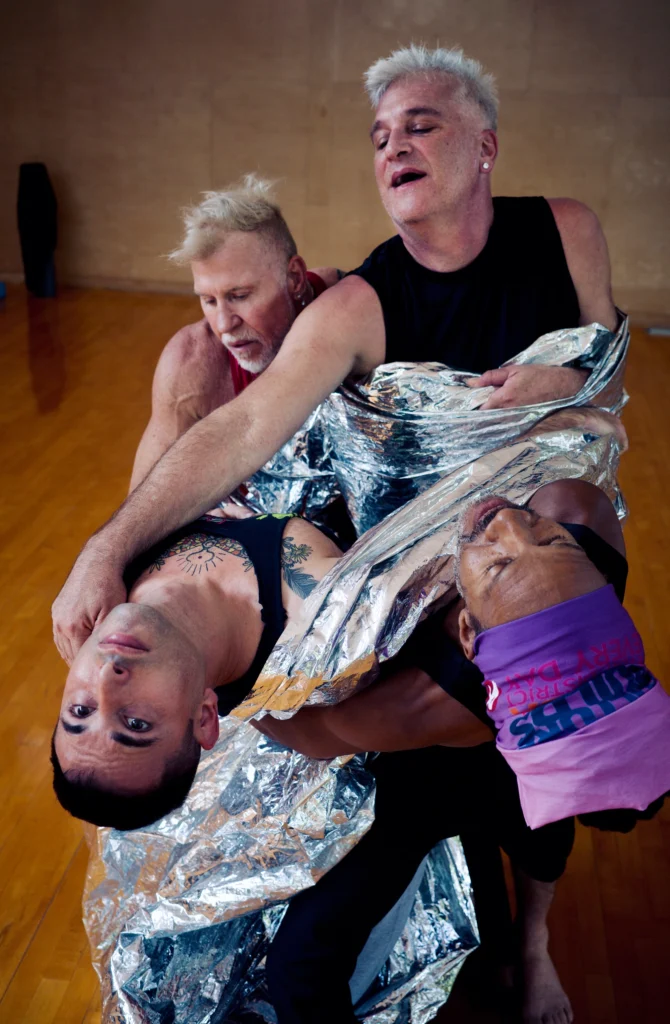 Four male-presenting dancers cluster together, wrapped up in shiny, metallic fabric. Two bend backward together, while the other two reach forward to support them.