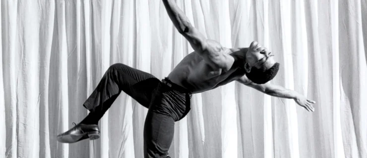 A black and white archival photo of a young Alvin Ailey, balanced on forced arch as he extends his upstage leg in parallel attitude and leans back so his head is parallel to the floor. His arms are extended in opposition; he is shirtless and wears dark pants and shiny shoes.