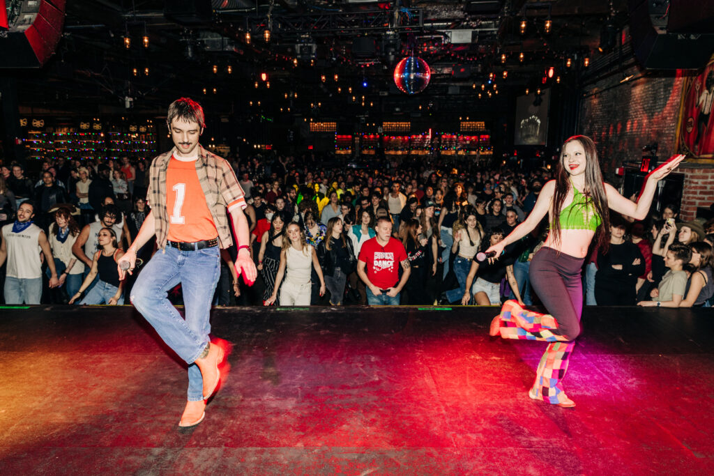 two dancers on stage leading a crowd behind them in a line dance