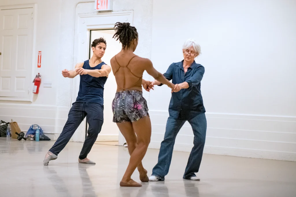 Twyla Tharp holds the hands of a dancer, the two sketching a tendu front with bent supporting leg. Another dancer watches, imitating Tharp's movements behind them.