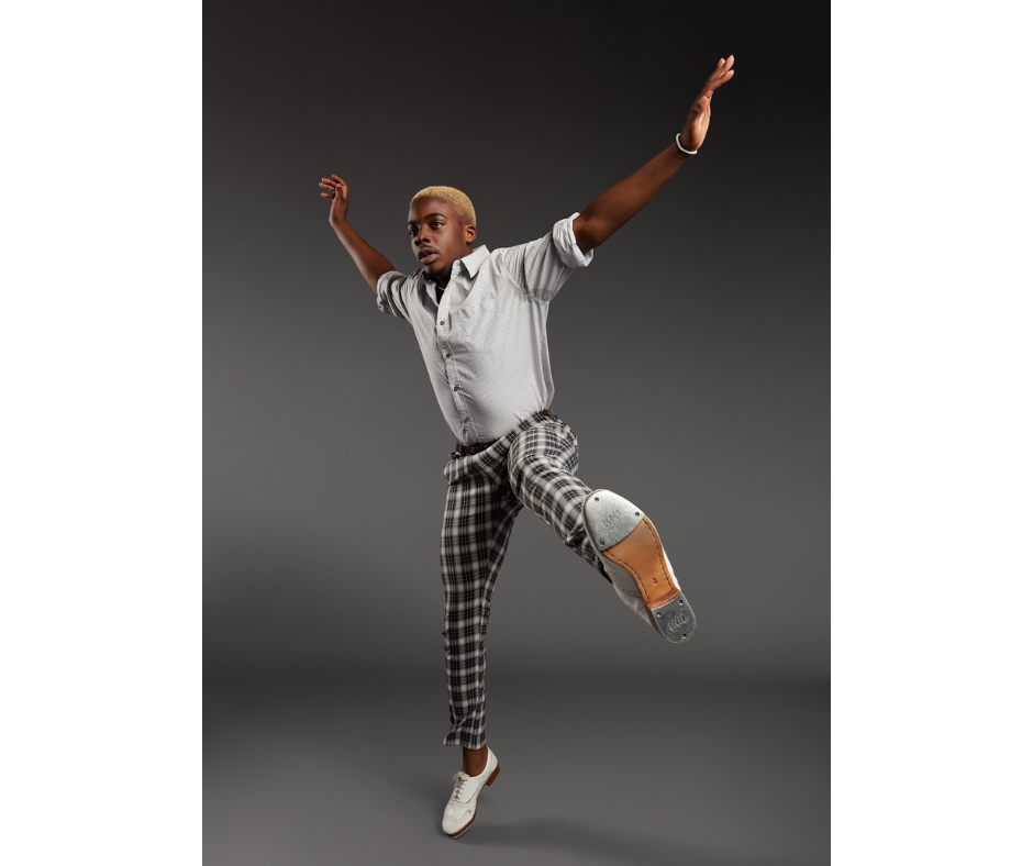 a male tap dancer wearing plaid pants and a button down shirt jumping up with his limbs spread wide