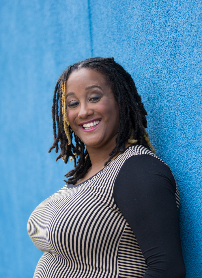 a headshot of Kayla Hamilton. She poses in profile in front of a light blue background. Kayla is a dark brown-skinned Black woman. She wears a long sleeve black & white striped shirt & her dreadlocks are down. She has a beautiful glowing smile.