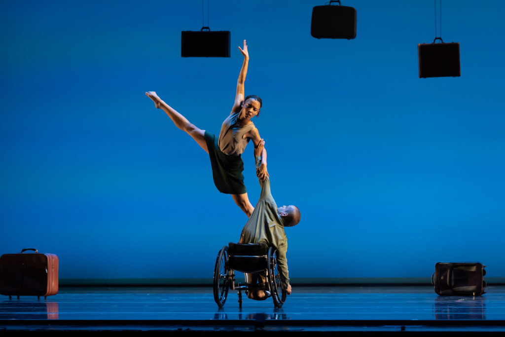 JanpiStar holds a side lunge in their wheelchair; Alaja places one foot on Janpi's knee and soars through space with their other leg lifted in an upward extension. Luggage hangs from the ceiling above the dancers.