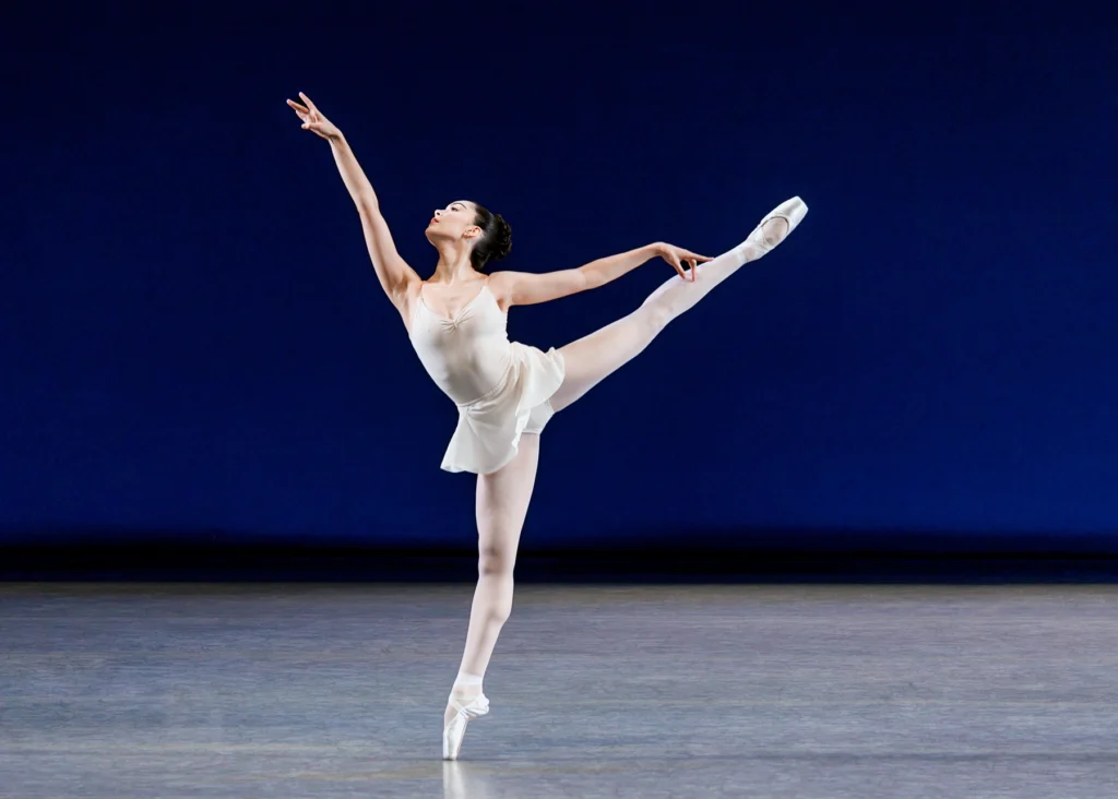 Mira Nadon is alone onstage as she balances in a high first arabesque, front arm exaggeratedly high and chin tipped up to follow it. She wears a white leotard and skirt over pale pink tights and pointe shoes.