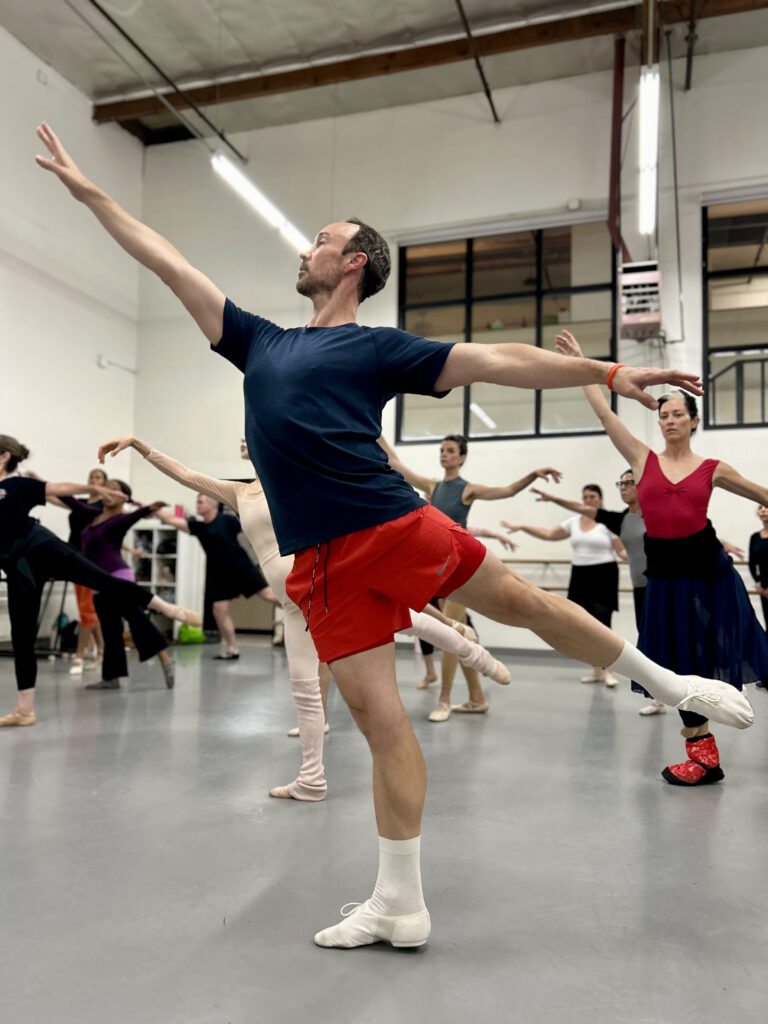 a male ballet teacher in first arabesque while leading a large group of dancers in the studio