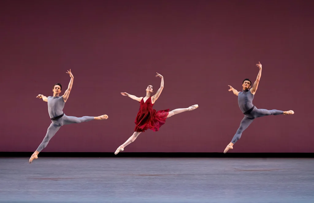 Mira Nadon is caught midair between two male dancers, all doing identical sissonnes with their back arms in high fifth. Nadon wears a flowing red dresses, pink tights, and pointe shoes; the men are in grey unitards with black waistbands.