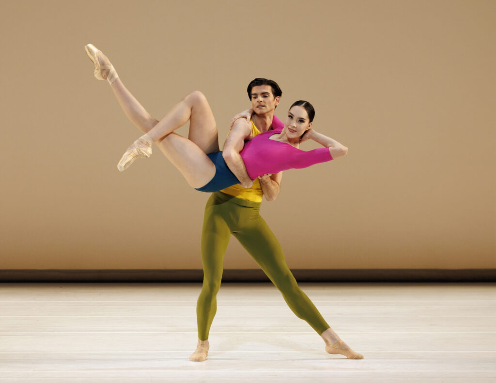 Mira Nadon wears a mischievous smile as she is swung into the air by her partner. She places one arm around his neck, while the other hand rests behind her head as though reclining on a couch. One leg is extended at an angle upwards, while the other pointed foot crosses daintily over that knee. She wears a color-blocked leotard in hot pink and navy blue, no tights, and pink pointe shoes.