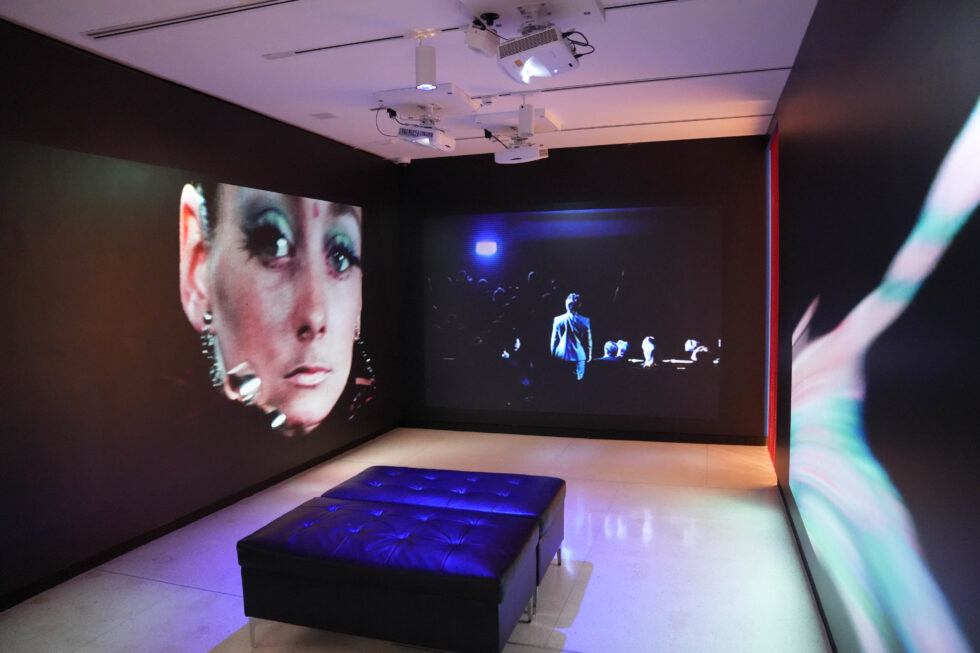 A photo of a small room with black walls hung with projector screens. The screens feature images from the psychedelic ballet "Astarte"—a close-up of a woman's face, a group shot.