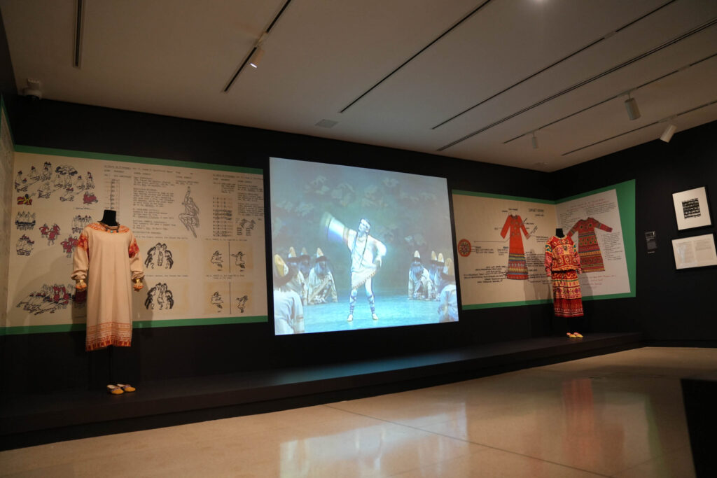 A photograph of a museum room, with a large screen in the center playing a scene from "Rite of Spring." Brightly-colored costumes from the production are displayed on either side of the screen.