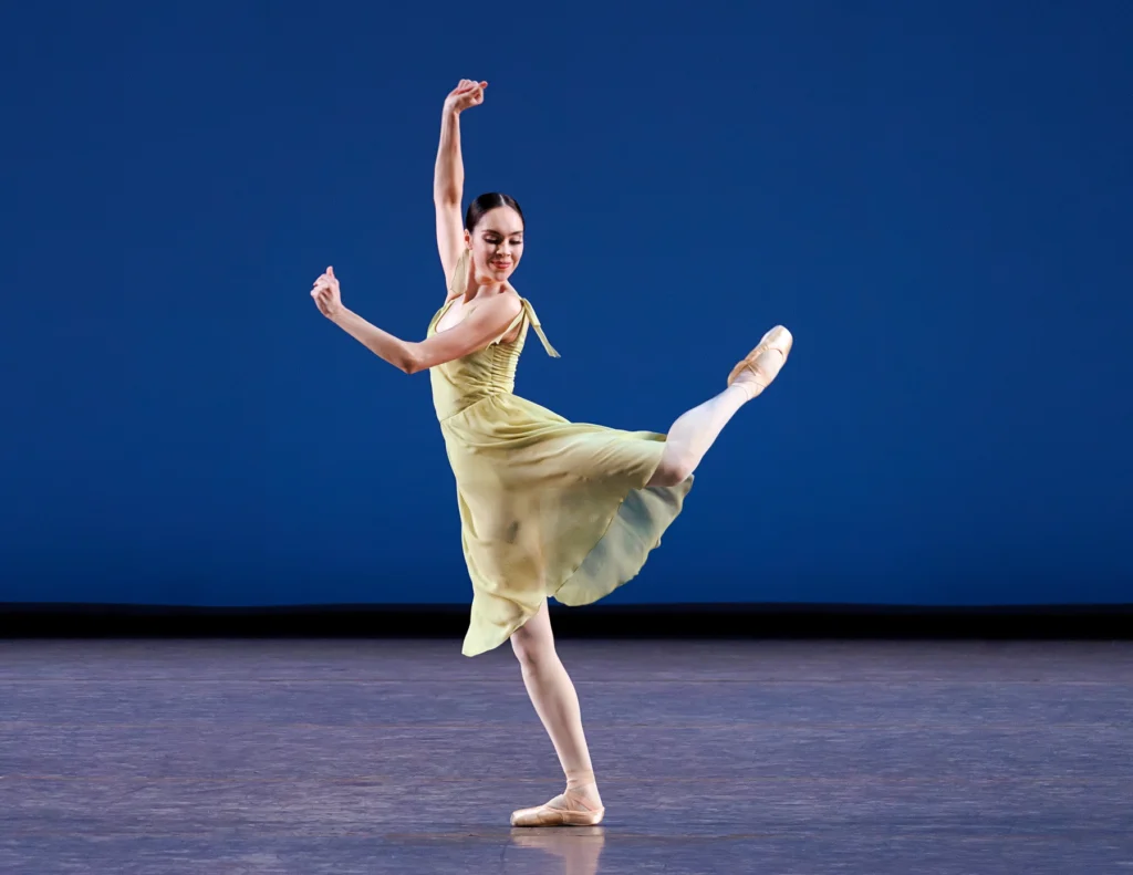 Nadon glances over her shoulder with a smirk as she flicks a leg into a loose back attitude. She is on flat, standing knee slightly bent, arms raised in front of her chest and overhead with her fingers curled in, as though snapping. She wears a semi-transparent pale green dress over pink tights and pointe shoes.