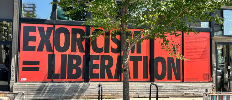 A view from the sidewalk of a building with glass plate windows. Across the windows, the words "Exorcism = Liberation" are printed in large black capital block letters on a red background.