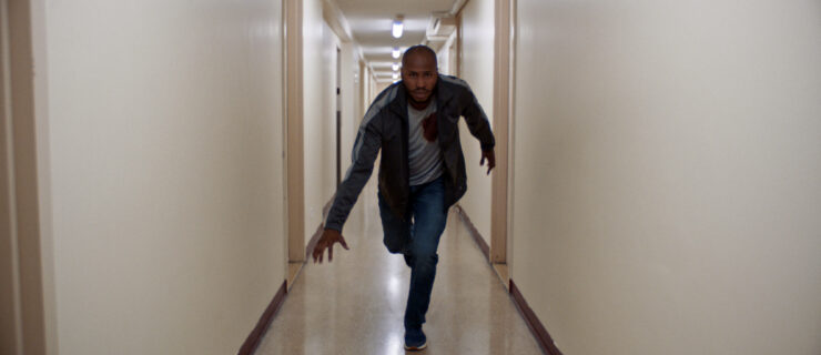 Bozeman—wearing jeans, a track jacket, and a blood-stained t-shirt—is pictured in a long white-walled hallway, lunging forward toward the camera.