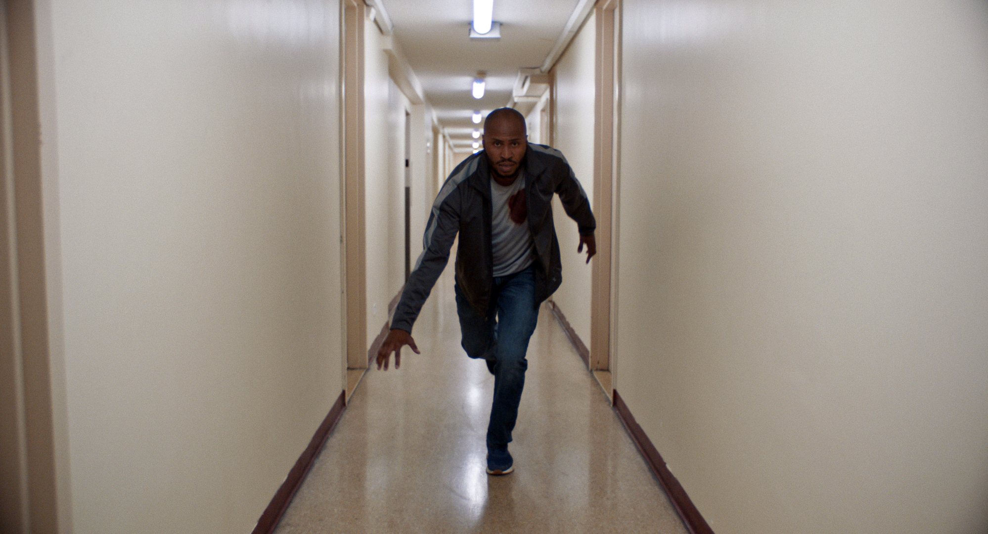 Bozeman—wearing jeans, a track jacket, and a blood-stained t-shirt—is pictured in a long white-walled hallway, lunging forward toward the camera.