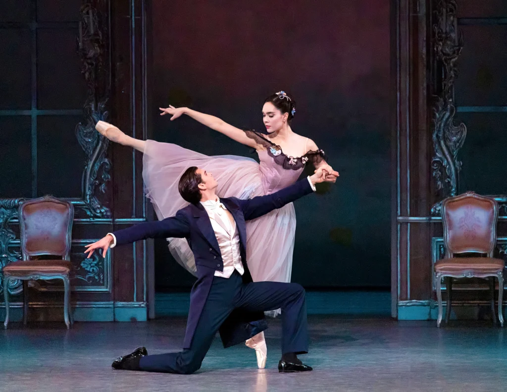 Nadon balances in first arabesque en pointe behind her partner, who kneels downstage of her and offers a hand for balance. She stares intently down at his face, expression almost severe; she wears a mauve romantic tutu that evokes a Victorian gown.