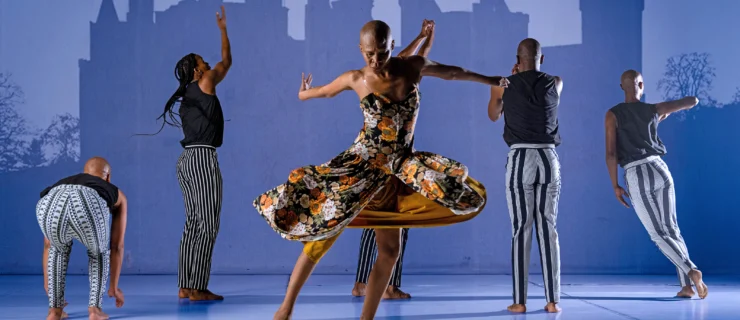 Dada Masilo spins at center stage, arms flung behind her, her floral dress flaring around her thighs. Her gaze is downward; her feet are barefoot. Upstage, five dancers in black and white face a shadowy outline of a building projected on the back wall.