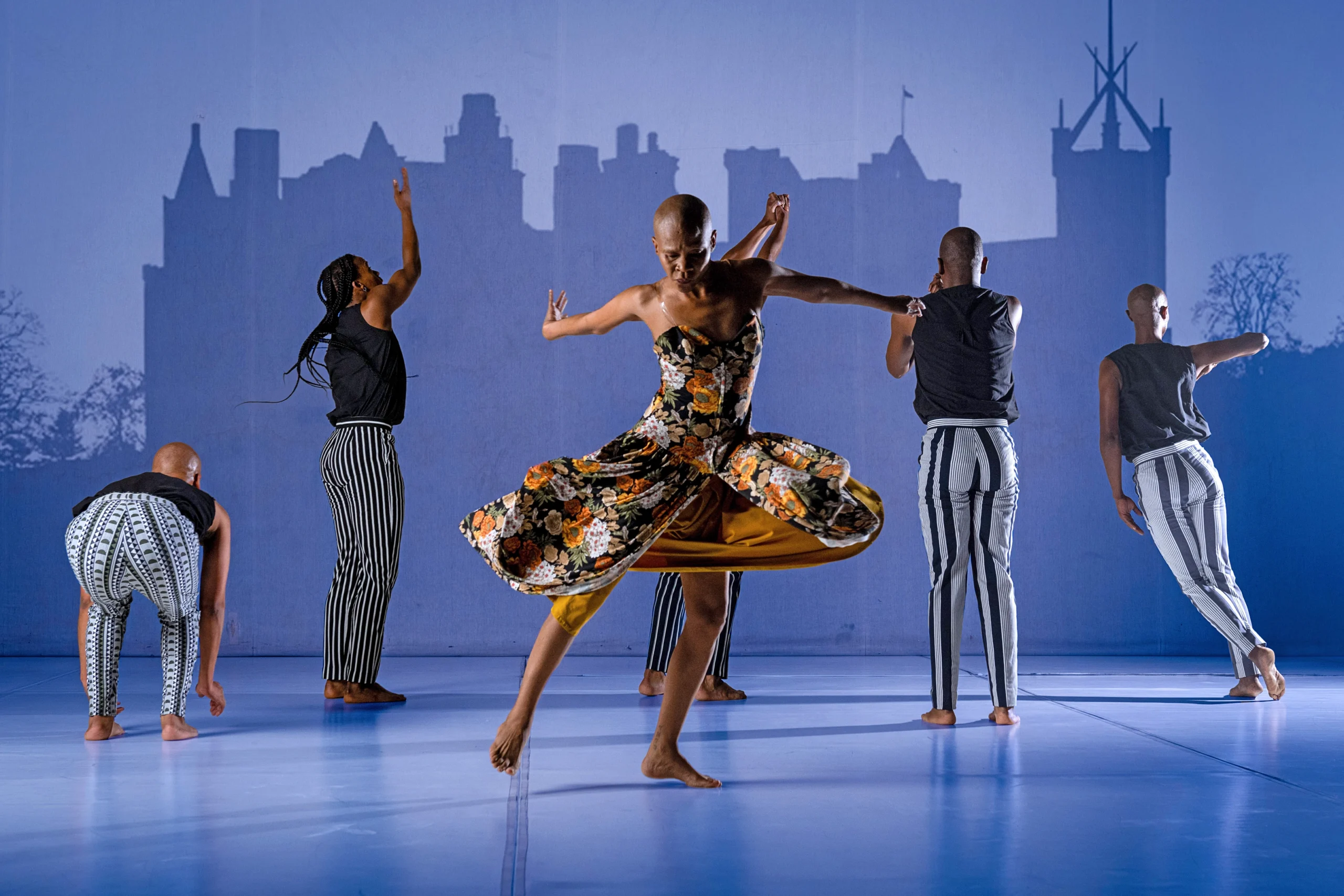 Dada Masilo spins at center stage, arms flung behind her, her floral dress flaring around her thighs. Her gaze is downward; her feet are barefoot. Upstage, five dancers in black and white face a shadowy outline of a building projected on the back wall.