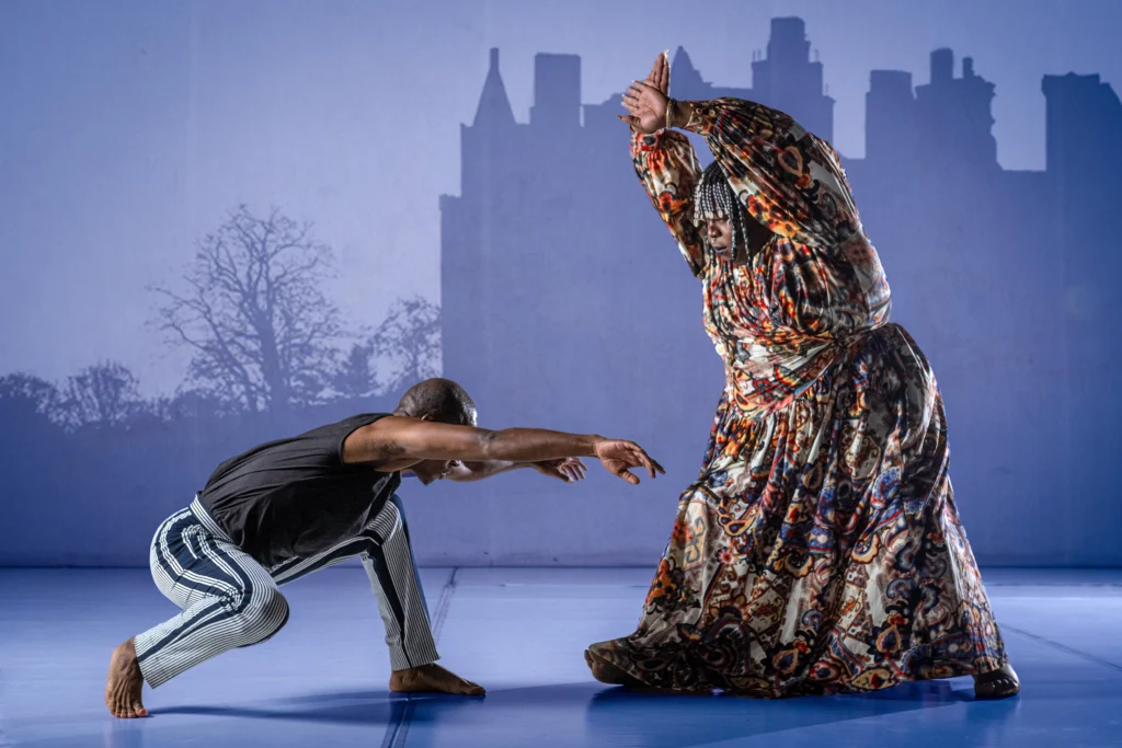 A dancer crouches low and folds over their front leg, arms outstretched parallel to the floor. Standing over them is a full-figured singer in a brightly patterned full-length dress; their arms raise overhead, palms facing out toward the crouching dancer. The backdrop is mauve with the silhouette of a castle and trees.