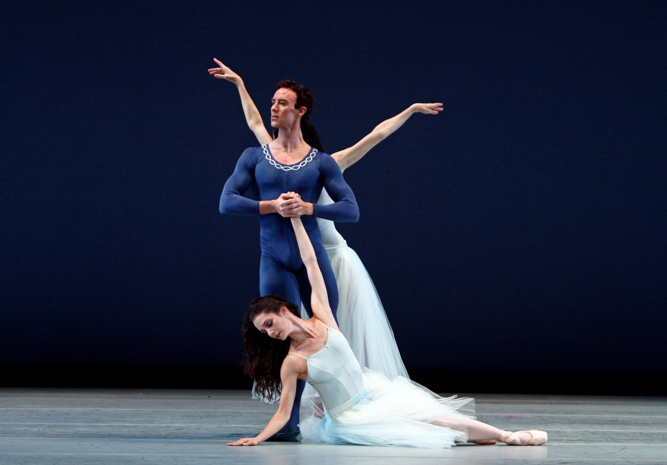 a male dancer holding the hand of a female dancer lying on the floor with another female dancer moving behind him