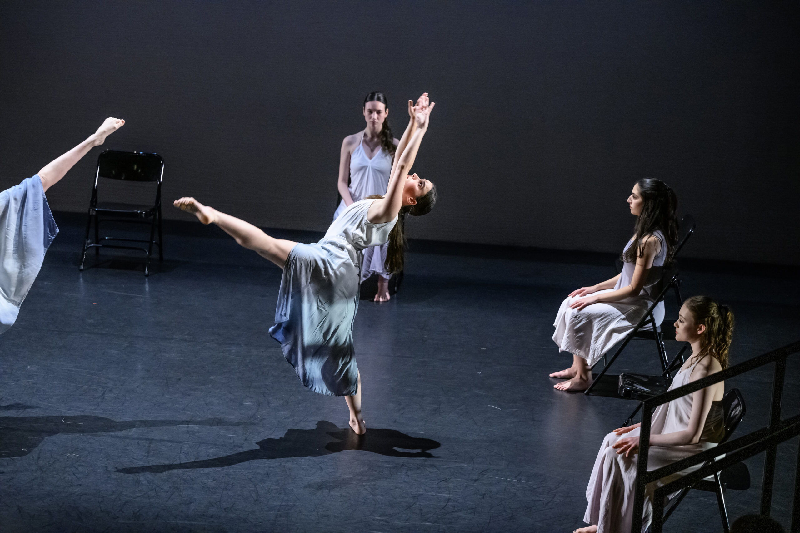 Four dancers on stage at Skidmore College. One dancer is extending her left leg and reaching up towards the ceiling. The other three are sitting in chairs watching.