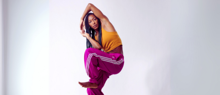 a female dancer wearing purple joggers and an orange tank posing in a contracted position with one leg lifted and her arms connected overhead