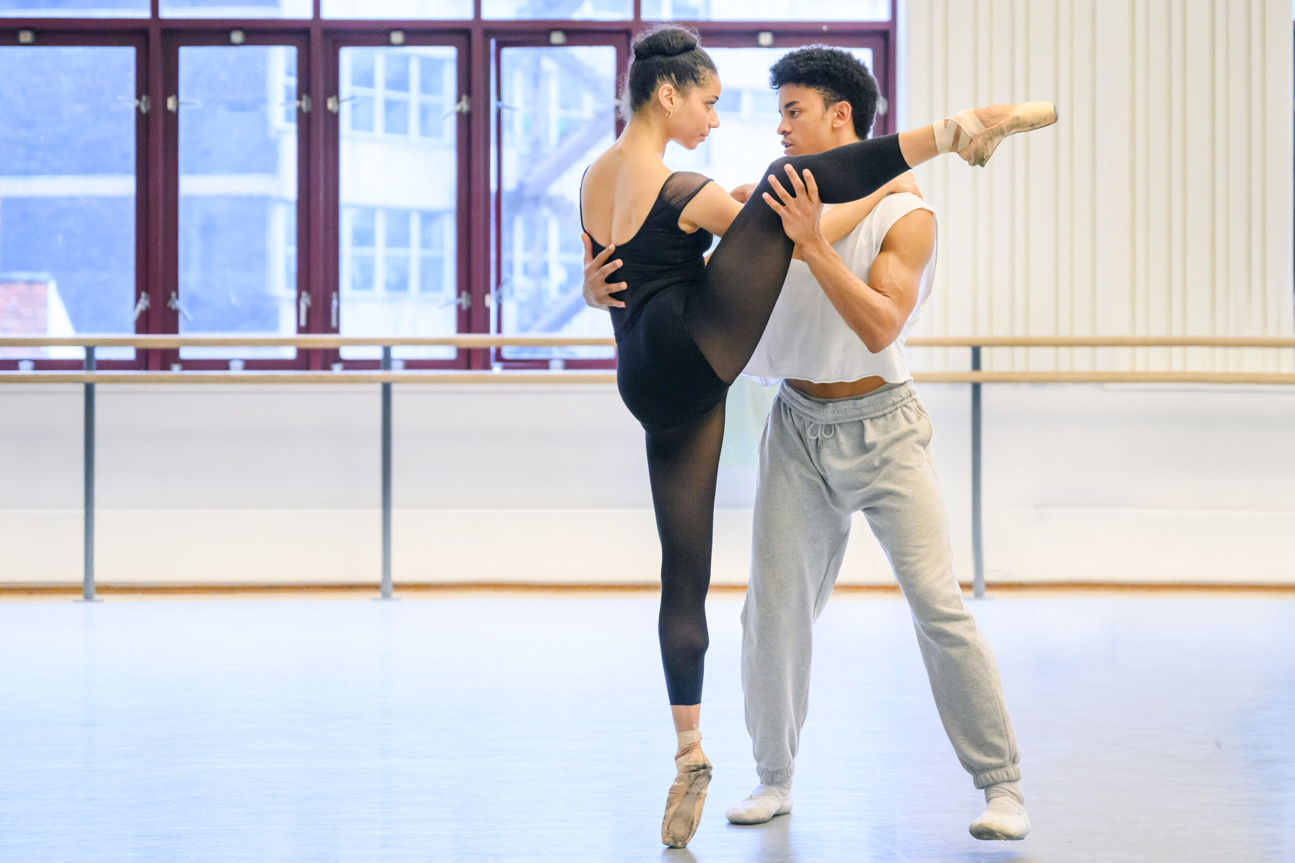 Two dancers in rehearsal clothes lock eyes with intensity. The woman in pointe shoes balances in a high side attitude; her partner holds her with one hand at her waist, the other on the outside of her knee.