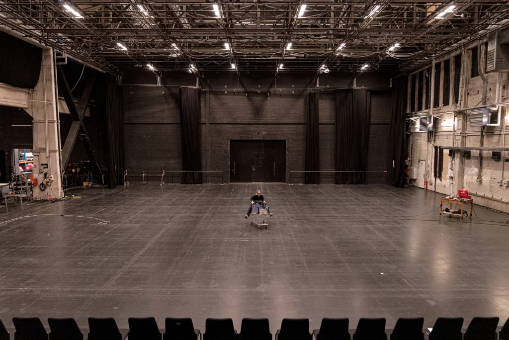 Oona Doherty rides a shopping cart, tiny in the midst of a cavernous theater space absent a back curtain or wings.