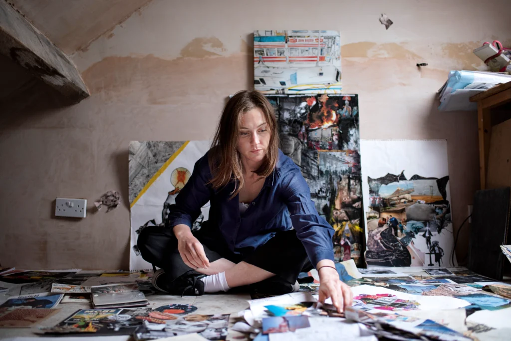 Oona Doherty purses her lips thoughtfully as she reaches to rearrange materials for a collage. She sits cross-legged on the floor, surrounded by colorful scraps. Her hair is brown and waves around her shoulders; she wears a navy tracksuit and sneakers.