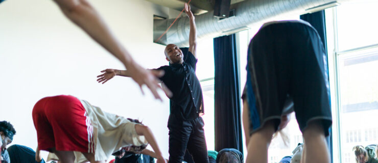 Shamel Pitts smiles widely as he moves in the center of a dance studio. He stretches one arm to the side and the other, slightly curved, overhead while around him students hang forward over their knees.