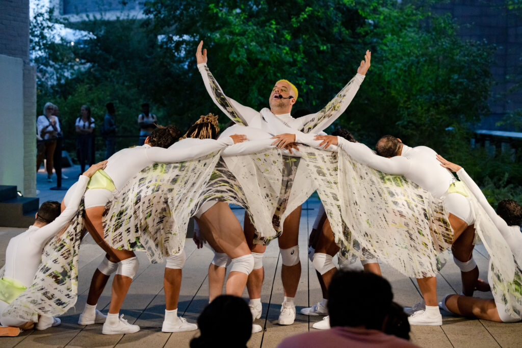 A group of dancer performing wearing long-sleeved white costumes with fabric attached to the arms. One dance is lifting his arms in the middle with the rest huddled around 