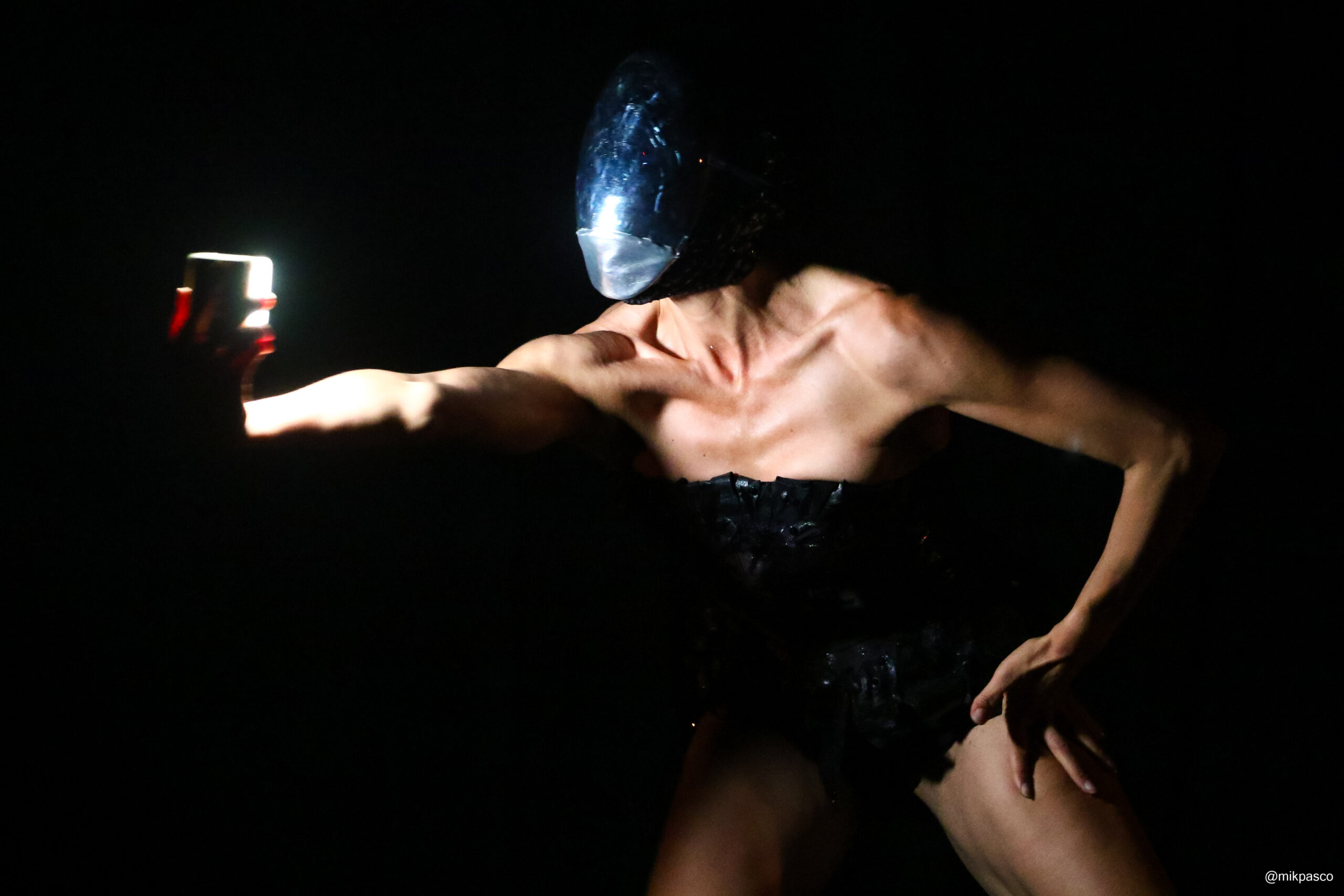 A dancer in a black costume and black mask covering her entire head stares into a bright light shining from her extended right hand. Her shoulders are bare.