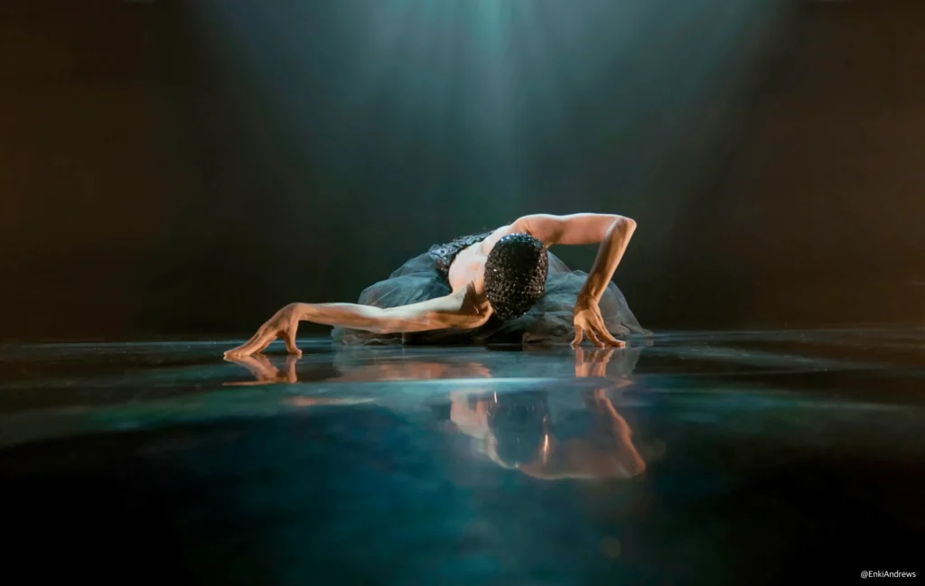 A kneeling dancer in a black costume with a black mask stretches on a reflective stage under a spotlight, with shoulders bare, extended arms, and head down.