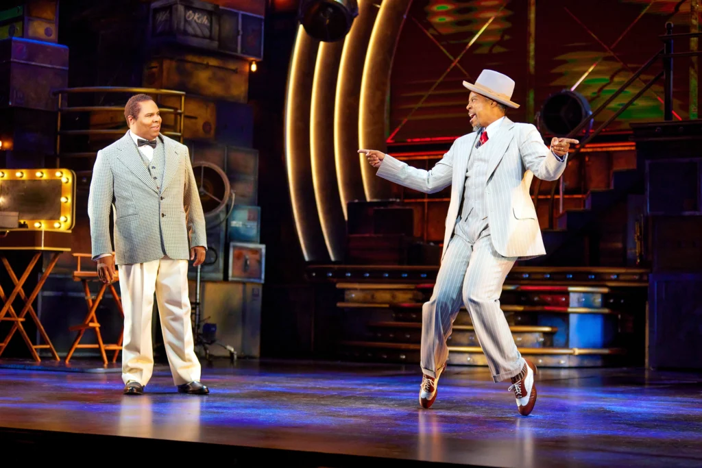 A man in a light-colored suit with a bow tie looks on as another man in a white suit and fedora balances on the tips of his tap shoes, knees knocking in, fingers pointing to either side.