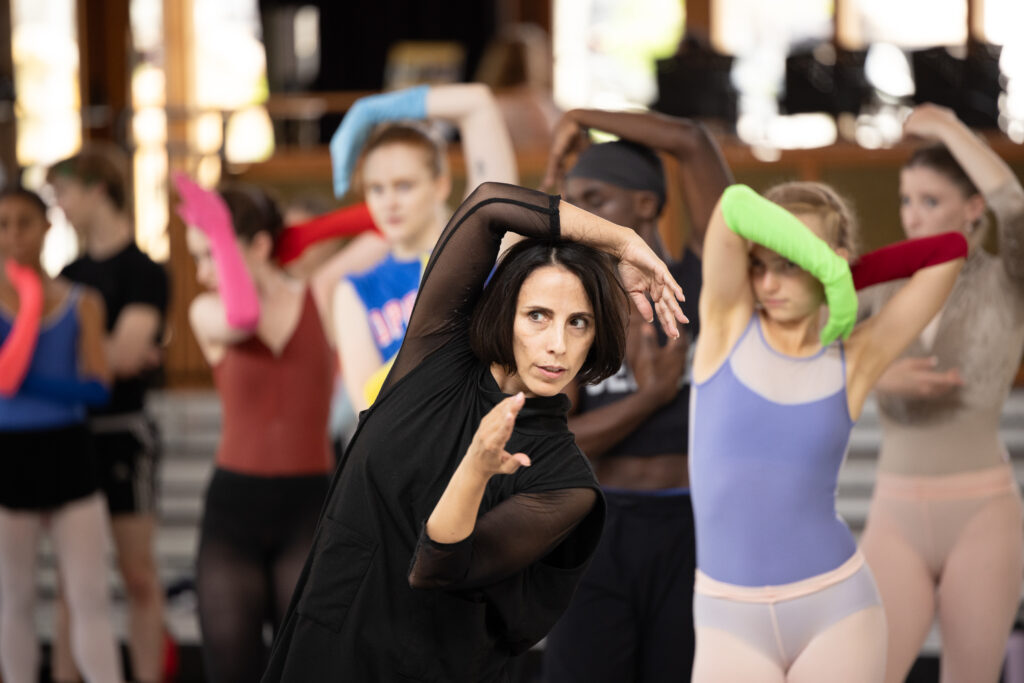 Annabelle Lopez Ochoa leading rehearsal wearing all black. She poses with her hands around her face and a group of dancers follow behind 