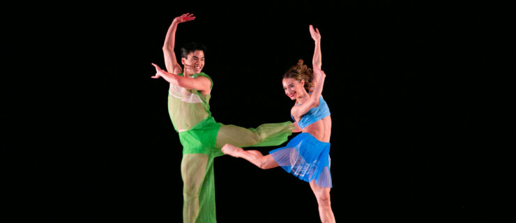 Austin Kelly and Lisa Borres dancing on stage wearing bright green and blue costumes. The two dancers are leaping in the air while throwing their arms to the side